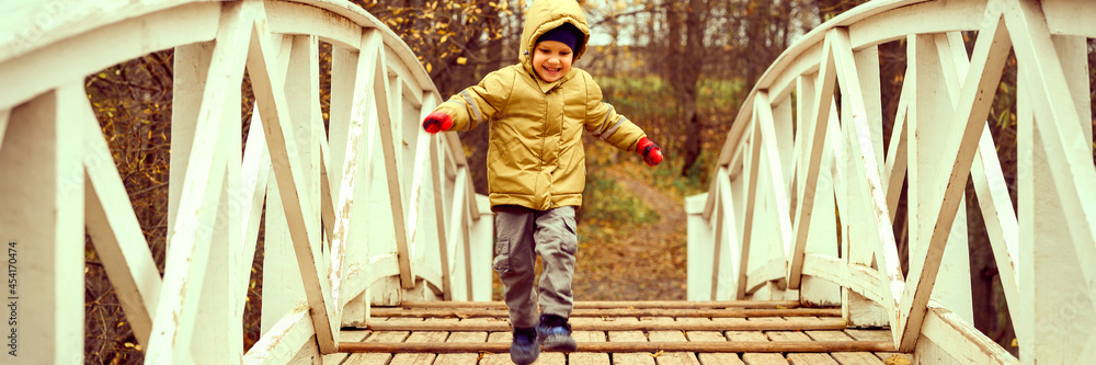 Φωτογραφία little happy four year old kid boy in autumn clothes runs and jumps on the humpback wooden bridge in the fall park