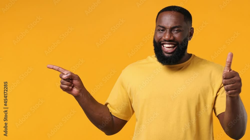 Young african american man 20s wears orange t-shirt pointing finger hand aside on workspace copy space mockup promo area show thumb up like gesture isolated on plain yellow background studio portrait Young african american man 20s wears orange t-shirt pointing finger hand aside on workspace copy space mockup promo area show thumb up like gesture isolated on plain yellow background studio portrait
