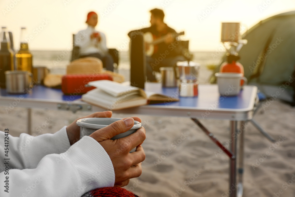 Obraz premium Friends resting near camping tent at beach, focus on woman with cup of hot drink