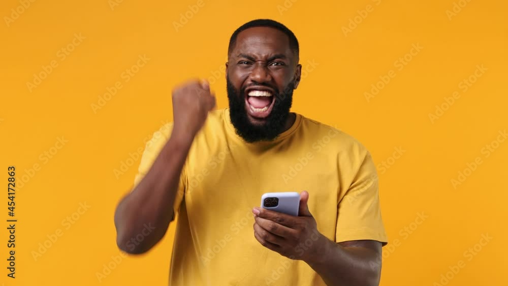 Young african american man 20s wears orange t-shirt hold use mobile cell phone typing say yes just found out great big win news doing winner gesture isolated on plain yellow background studio portrait Young african american man 20s wears orange t-shirt hold use mobile cell phone typing say yes just found out great big win news doing winner gesture isolated on plain yellow background studio portrait