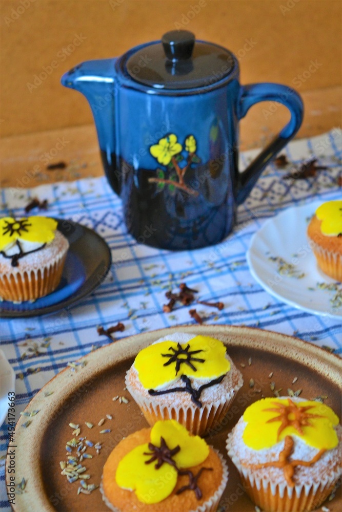 Fototapeta premium Blue coffee set with yellow flowers surrounded by muffins decorated with powdered sugar and icing in the shape of a yellow flower that matches those of the coffee set on a white and blue squares