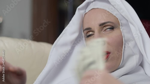 Close-up portrait of narcissistic Caucasian beautiful woman in nun costume counting dollars using money as fan. Headshot of seductive confident rich lady looking at camera smiling posing with cash photo