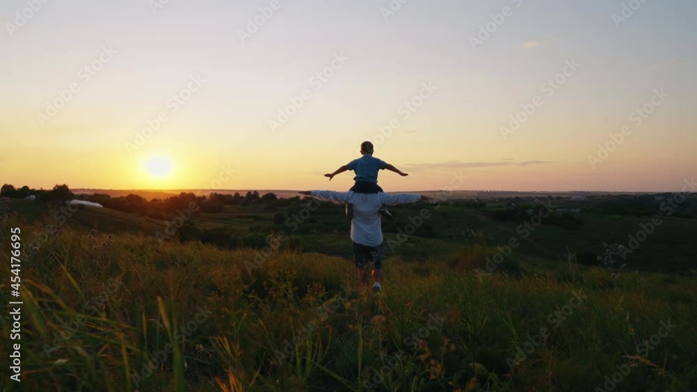 Father and son playing on grassy hill, little boy sitting on shoulders of parent, both spreading arms as if flying, country houses in the distance. Following shot loving family at sunset Father and son playing on grassy hill, little boy sitting on shoulders of parent, both spreading arms as if flying, country houses in the distance. Following shot loving family at sunset
