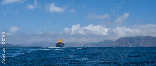Small boat behind cruise ship near Santorini island in Aegea sea, Greece. Ferry sailing to port. Transportation by sea.