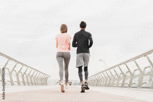 Fototapeta Naklejka Na Ścianę i Meble -  Young fit friends couple boyfriend and girlfriend athletes jogging running together in fitness outfits and trainers on city bridge outdoors.
