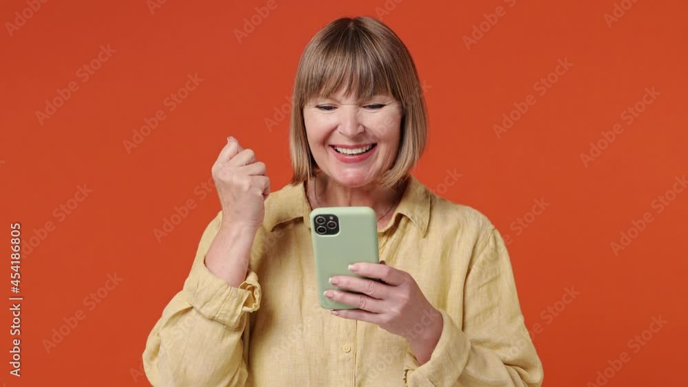 Happy mature elderly senior elegant lady woman 55 years old wear yellow shirt hold use mobile cell phone typing say wow yes just found out great big win news isolated on plain orange background studio Happy mature elderly senior elegant lady woman 55 years old wear yellow shirt hold use mobile cell phone typing say wow yes just found out great big win news isolated on plain orange background studio