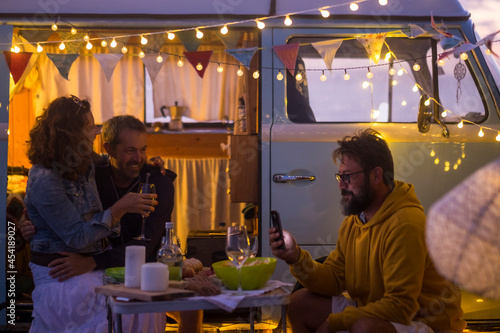Group of friends enjoy leisure night together with food and drins for dinner outside the van camper vehicle - travel and vacation summer lifestyle with men and woman having fun together