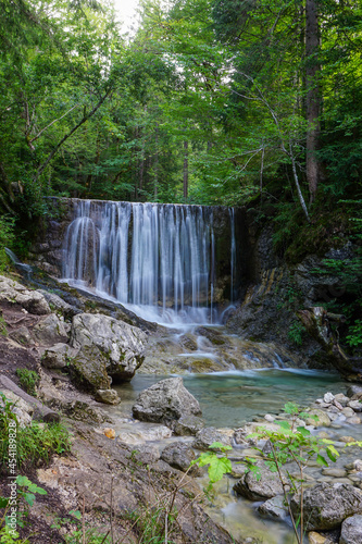 Ein Wasserfall fließt in einen Fluss. Im Vordergrund stehen große Steine. Oberhalb des Wasserfalls befindet sich eine weitere Ebene mit Bäumen.
