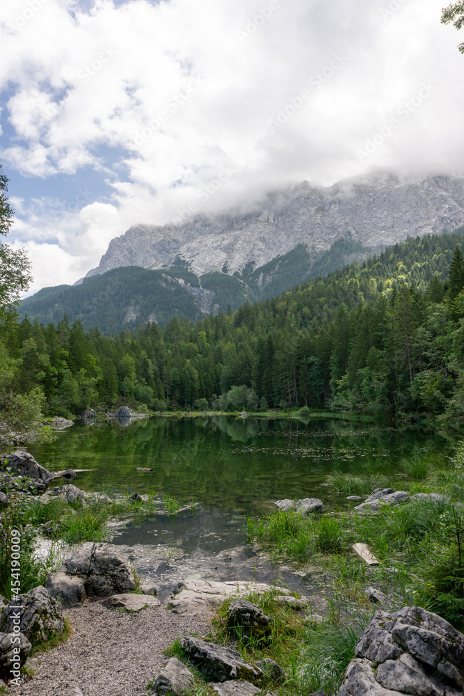 Obraz premium Ein kleiner grüner Teich, umgeben von einem Wald und Steinen. Im Hintergrund ist ein von Wolken umgebener Berg zu sehen. Die Bäume spiegeln sich im Teich.