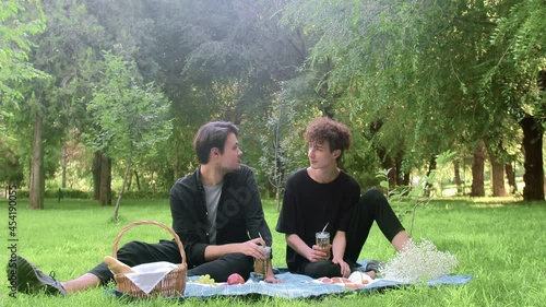 Two young guys sit on a picnic chatting and holding glasses of drinks in the summer