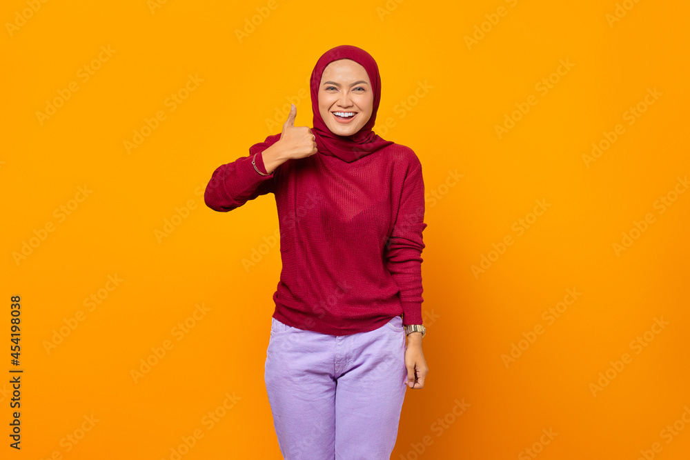 Happy Asian woman showing thumbs up and looking at camera over yellow background Happy Asian woman showing thumbs up and looking at camera over yellow background