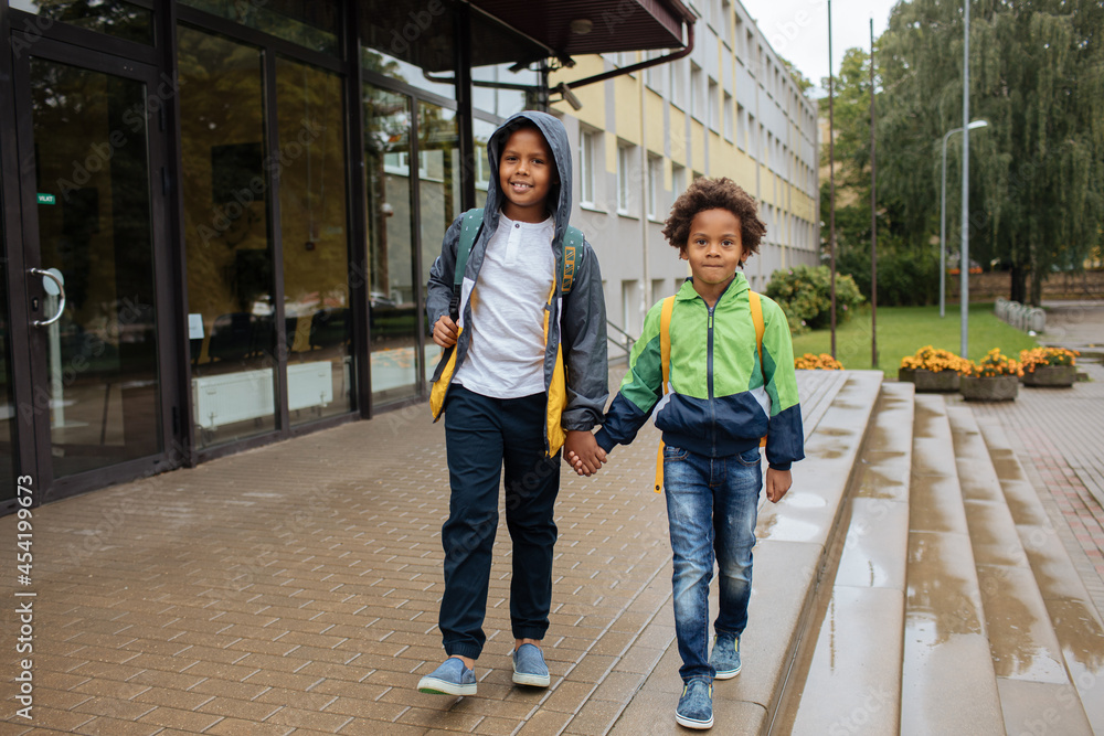 Little boys with back packs. Two African American boys next to the school. Back to the school concept. Kids standing outside and happy to meet each others. Happy black children. First day of school