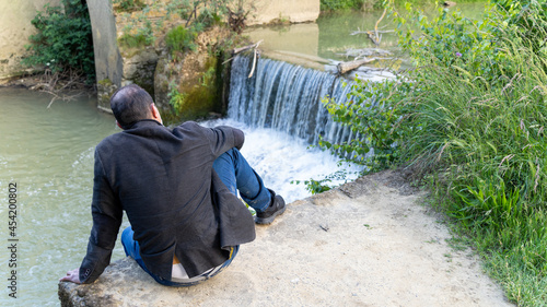 Young man from behind, sitting at the edge of a waterfall, on a late spring day 