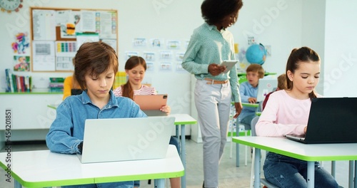 Children learning at primary school. Portrait of Caucasian schoolgirl and schoolboy typing on laptop surfing internet at computer science class African American female teacher tapping on tablet behind © VAKSMANV