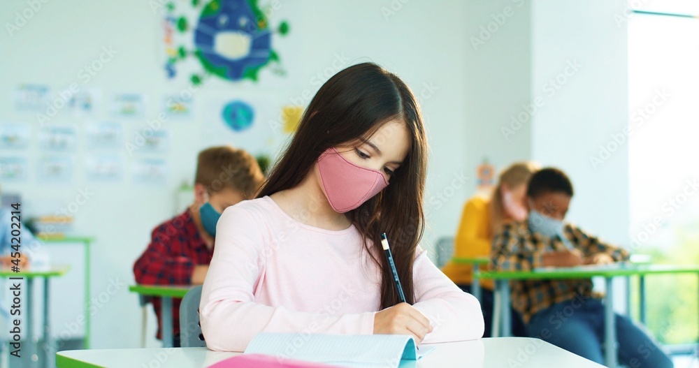 Portrait of beautiful concentrated schoolgirl in face mask studying writing at school while sitting at desk and listening to teacher. Portrait of beautiful concentrated schoolgirl in face mask studying writing at school while sitting at desk and listening to teacher.