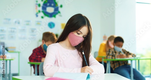 Portrait of beautiful concentrated schoolgirl in face mask studying writing at school while sitting at desk and listening to teacher.