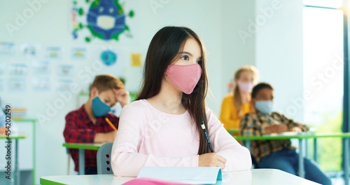 Close up portrait of Caucasian little pretty schoolgirl in mask writing sitting at desk at school and raising hand. Mixed-race kids in classroom on background. Coronavirus pandemic, primary education