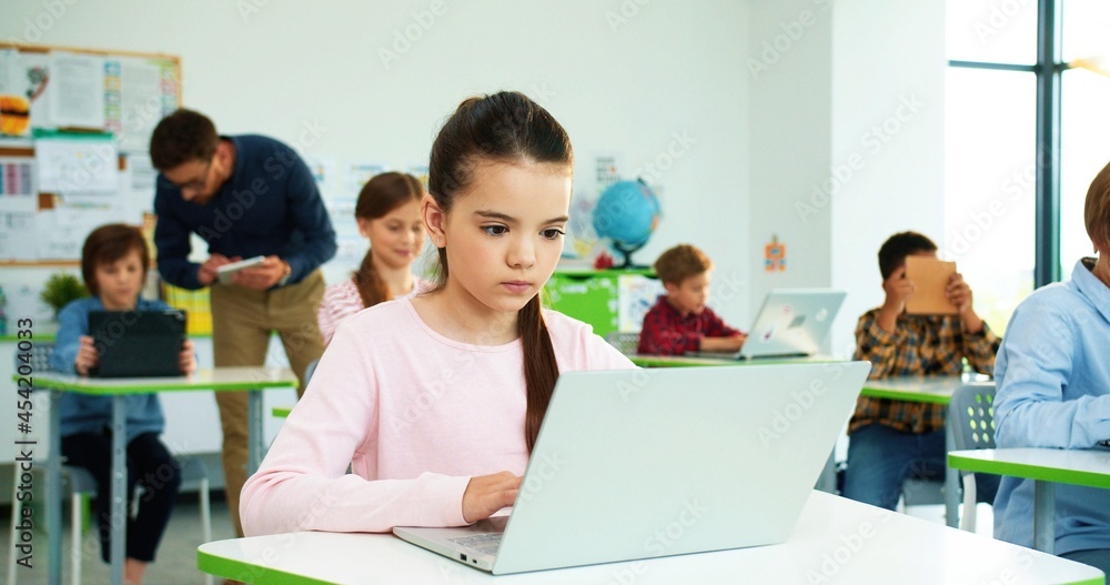 Close up of concentrated Caucasian little girl sitting at desk at school class typing on laptop Children studying in classroom using digital gadgets, computer science class modern elementary education Close up of concentrated Caucasian little girl sitting at desk at school class typing on laptop Children studying in classroom using digital gadgets, computer science class modern elementary education