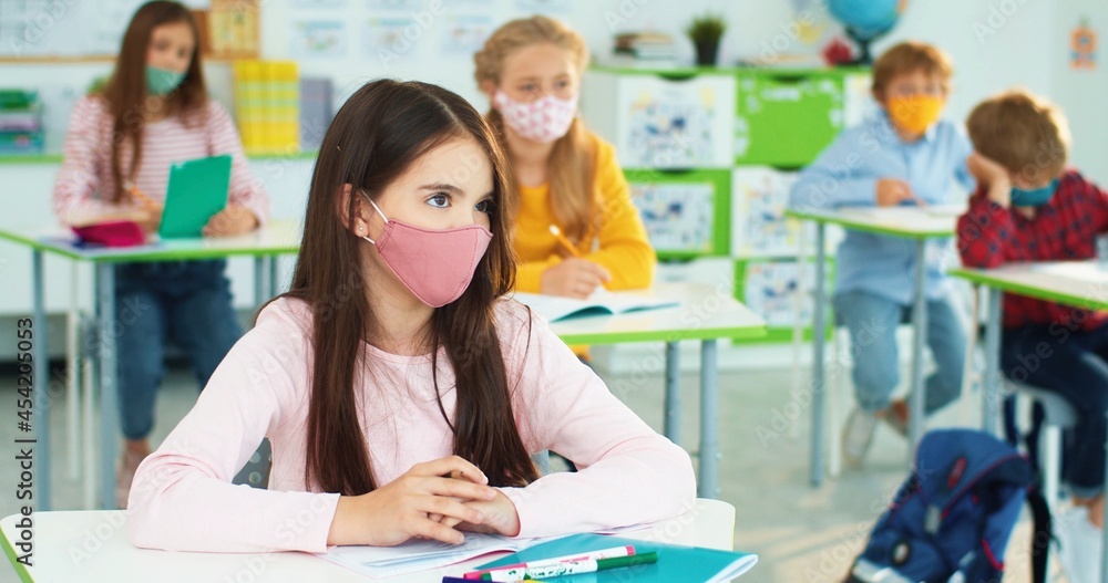 Close up portrait of Caucasian little bright schoolgirl in mask sitting at desk in classroom at lesson and listening to teacher. Diverse children studying, coronavirus quarantine, elementary school Close up portrait of Caucasian little bright schoolgirl in mask sitting at desk in classroom at lesson and listening to teacher. Diverse children studying, coronavirus quarantine, elementary school