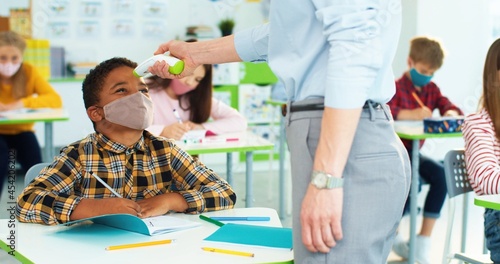 Close up portrait of female teacher's hand measuring temperature of small African American happy boy junior student in face mask sitting at desk using infrared digital thermometer, coronavirus