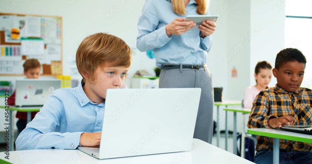 Close up of Caucasian little primary school student sitting at desk in classroom typing on laptop at lesson, female teacher walking behind using tablet. Mixed-race pupils learning at computer class Close up of Caucasian little primary school student sitting at desk in classroom typing on laptop at lesson, female teacher walking behind using tablet. Mixed-race pupils learning at computer class