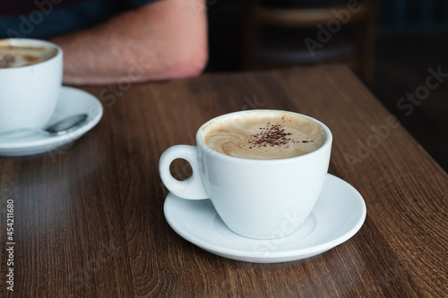 Chocolate sprinkles on the froth of a cappuccino coffee in a white cup and saucer on a dark brown wooden table. A second cup the arm of a man can be seen.