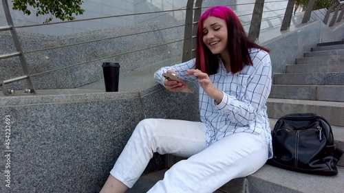 Happy beautiful red-haired student girl laughs heartily chatting on a smartphone in social networks sitting on the steps in the city. High quality 4k footage photo