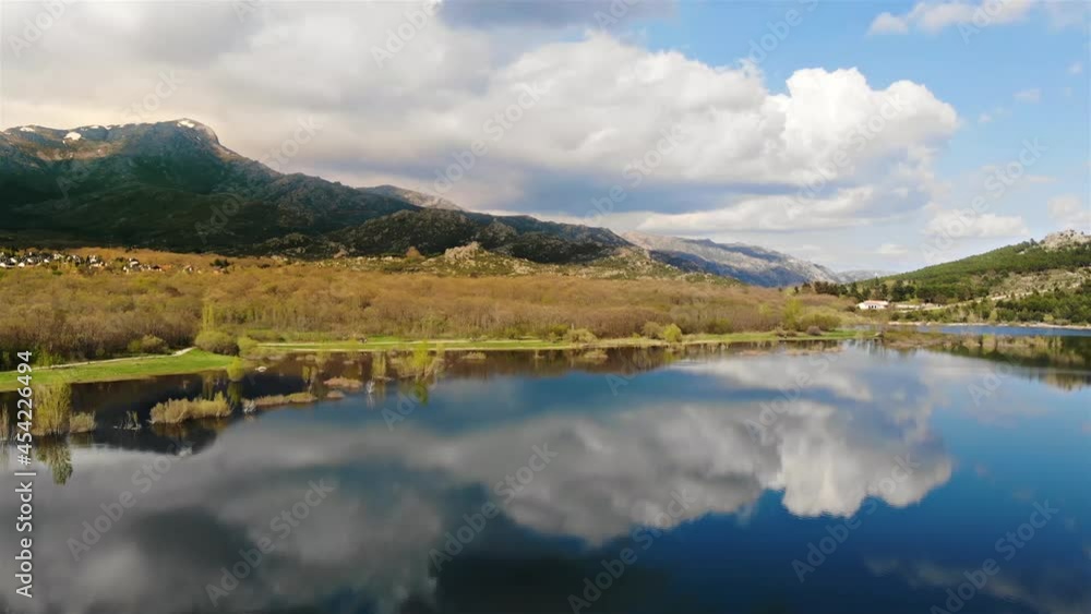 Long panning shot at a lake revealing snowed mountains and cosy village and a wide view of the clouds and the blue sky reflected on the water surface. Long panning shot at a lake revealing snowed mountains and cosy village and a wide view of the clouds and the blue sky reflected on the water surface.