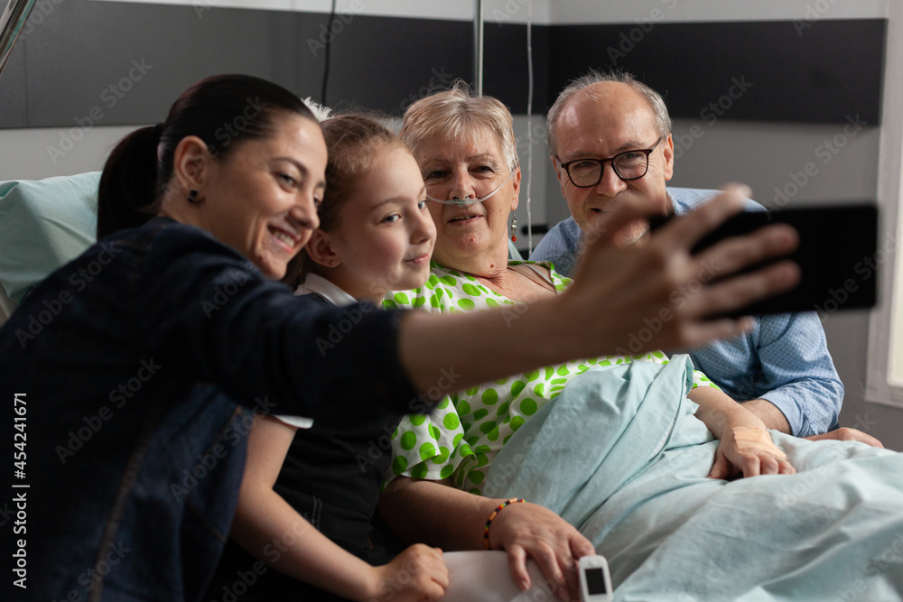 Family taking selfie with sick retired grandmother during medical recovery using modern smartphone in hospital ward supporting her after medicine surgery. Senior woman patient resting in bed