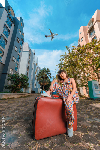 A cute adorable Asian girl about 6years old wearing a vintage dress. An active kid poses for a photo with an old red vintage suitcase alone with a large plane flying in the background. photo