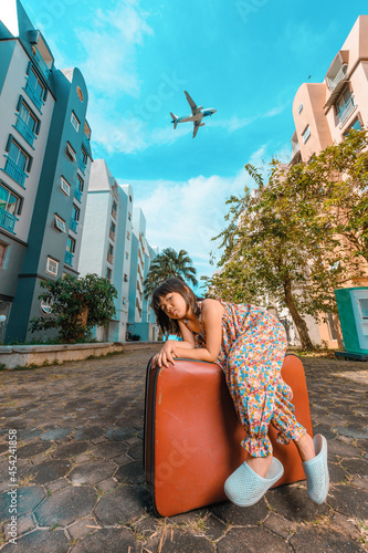 A cute adorable Asian girl about 6years old wearing a vintage dress. An active kid poses for a photo with an old red vintage suitcase alone with a large plane flying in the background. photo