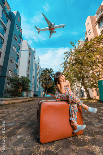 A cute adorable Asian girl about 6years old wearing a vintage dress. An active kid poses for a photo with an old red vintage suitcase alone with a large plane flying in the background. photo