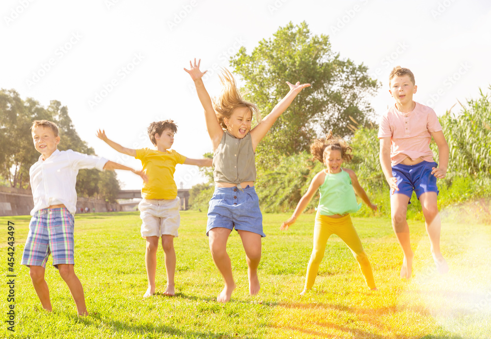 Fototapeta premium Positive kids jumping and raising hands up on green field.