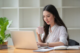 Photo of positive Asian woman holds a cup of coffee imagines plan holiday in office.