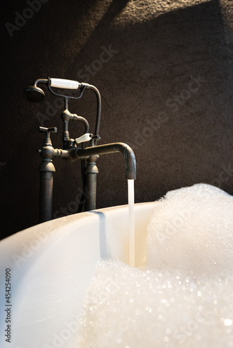 Water flowing from vintage designed tap into a bathtub with fully bubble foam, cleaning and relaxaing spa concept. Interior object photo. Close-up and selective focus in lowlight condition. photo