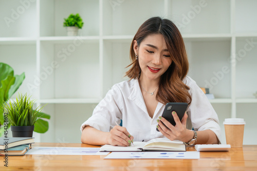 Confident Asian woman talking on her mobile phone and using laptop while sitting at desk and working from home. Home office. © PaeGAG