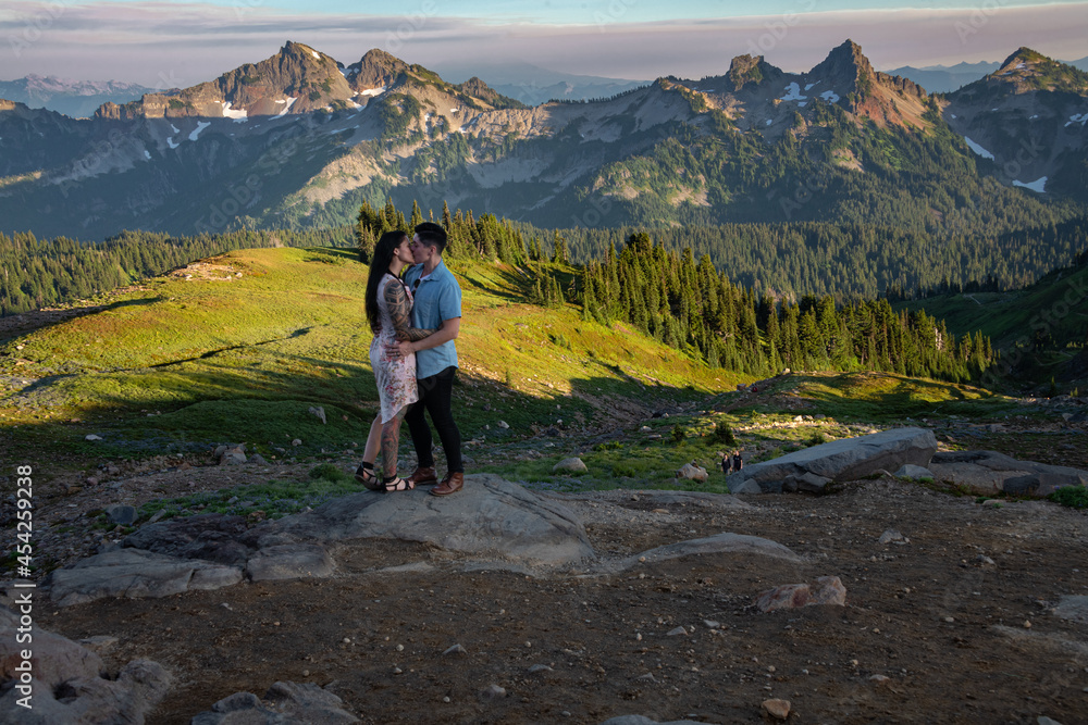 View of a couple kissing in front of mountains in Mount Rainier National Park during sunset. The landscape has rolling hills and several mountain peaks. 