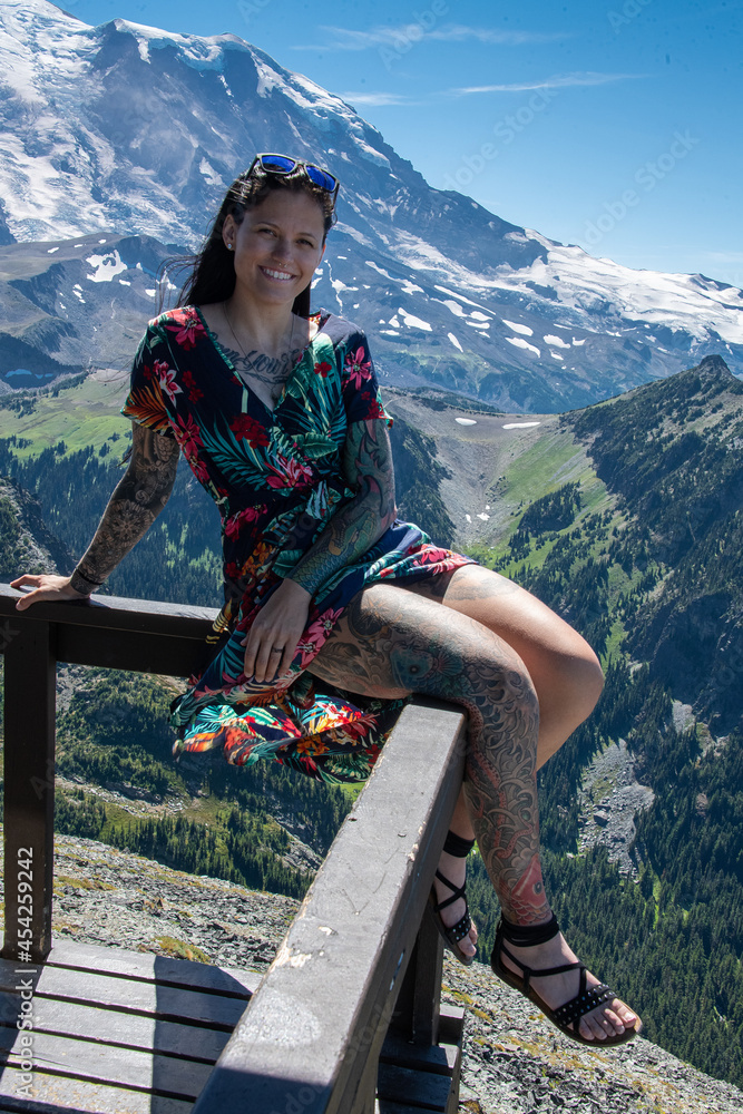 Naklejka premium View of a brunette young woman on the Mouunt Fremont Fire Lookout in Mount Rainier National Park. The peak of the mountain is covered in snow and below is fields of green grass.