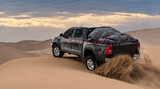 a black pickup truck is going up from a sand dune and splashing sands on air and around in dasht e lut or sahara desert with cloudy sky