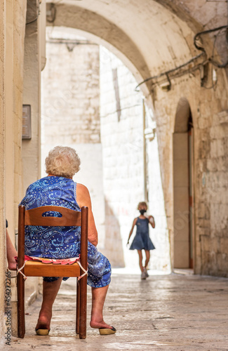 Fototapeta Naklejka Na Ścianę i Meble -  Grandmother waiting for her granddaughter on the doorstep in the narrow streets of Bari old town, Puglia, Italy, vertical