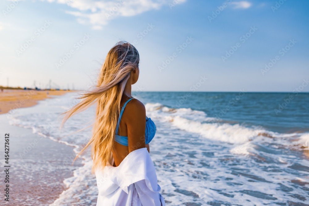 Naklejka premium A slender girl in a gentle blue swimsuit and shirt walks along the sandy beach near the blue sea