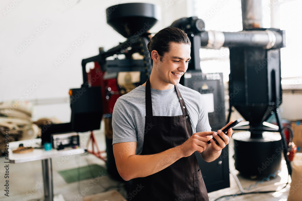 Portrait of handsome modern man wearing apron use phone while standing at coffee roasting machine in local roastery Portrait of handsome modern man wearing apron use phone while standing at coffee roasting machine in local roastery
