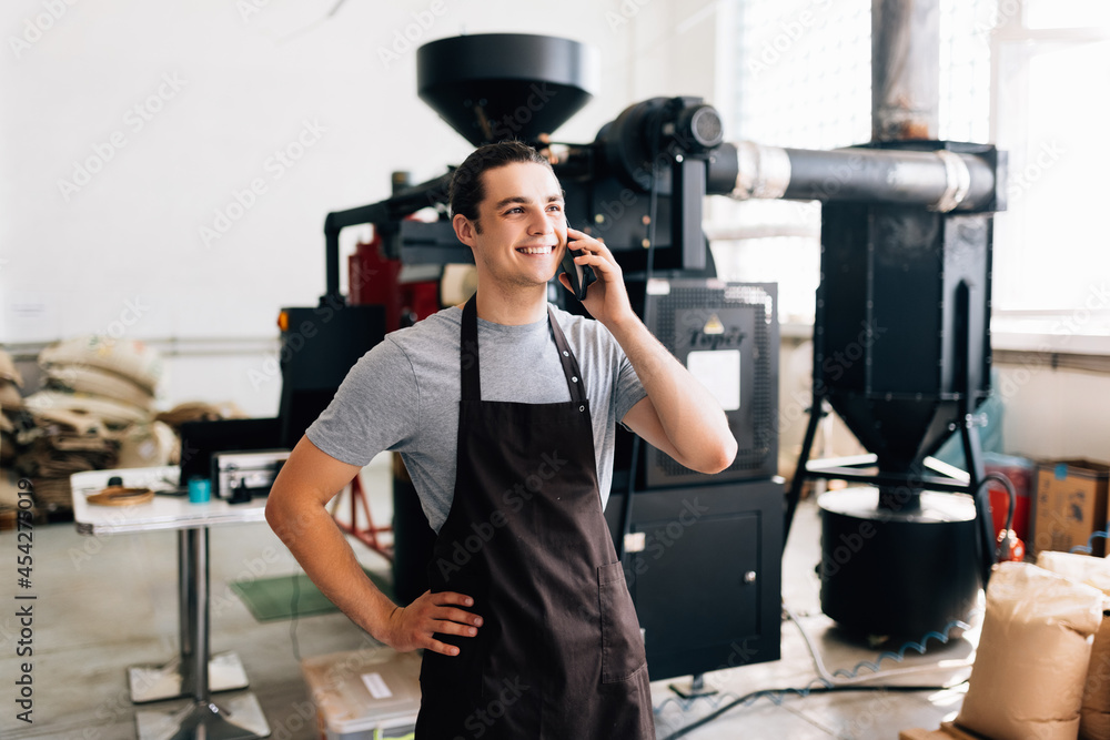 Portrait of handsome man wearing apron talk phone while standing at coffee roasting machine in local roastery Portrait of handsome man wearing apron talk phone while standing at coffee roasting machine in local roastery