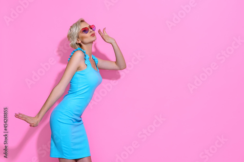 Portrait of beautiful blonde woman in blue dress with sunglasses posing on pink studio background