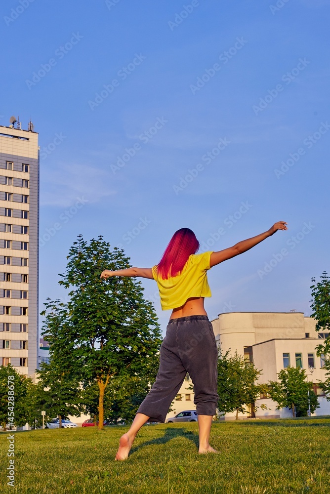 young woman with pink hair in a yellow t-shirt is dancing in a city park turned her back