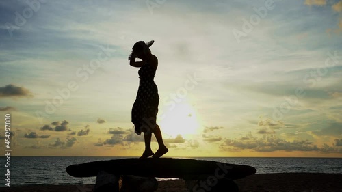Side view of short hair Asian woman silhouette in a beach dress and straw hat enjoy balancing walking and jump on a beach rocks with a calm sea and the evening sun in beautiful sky in the background. photo