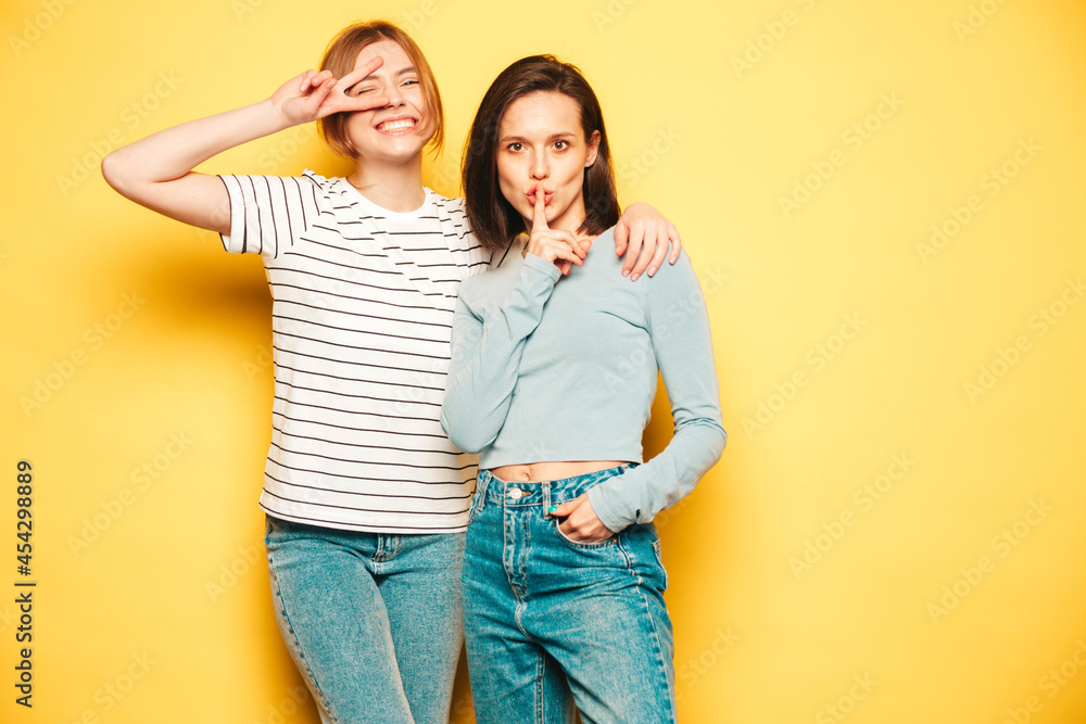 Two young beautiful smiling hipster female in trendy summer white t-shirt and jeans clothes.Sexy carefree women posing near yellow wall in studio.Positive and cheerful models Two young beautiful smiling hipster female in trendy summer white t-shirt and jeans clothes.Sexy carefree women posing near yellow wall in studio.Positive and cheerful models