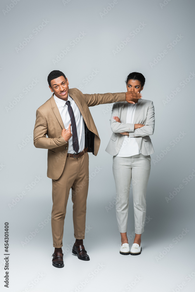 Fototapeta premium arrogant african american businessman covering mouth of businesswoman while pointing at himself on grey
