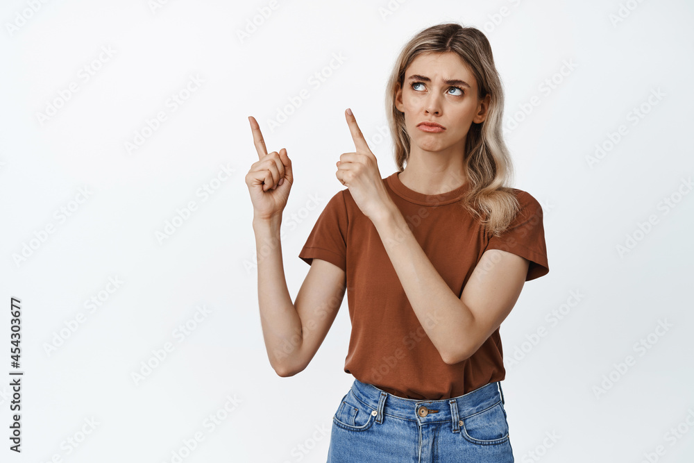 Sad and gloomy girl pointing fingers at upper left corner, whining and grimacing, standing against white background Sad and gloomy girl pointing fingers at upper left corner, whining and grimacing, standing against white background