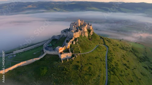 Aerial view of Spiš Castle, fog flows around the castle, lit by morning sun. Extensive castle ruins, standing on a 200m high white rock above the valley against mountains in background.  UNESCO site. photo